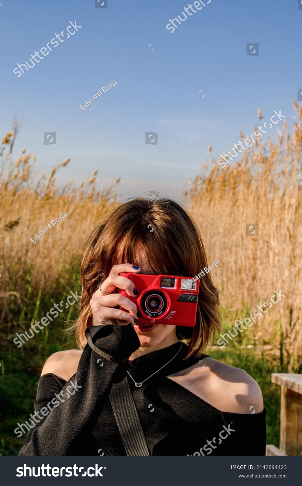 Vertical Photo of a Short-haired Girl Taking a Picture with a Red Retro Compact Camera in a Rice field with Tall Plants Background #2142894423