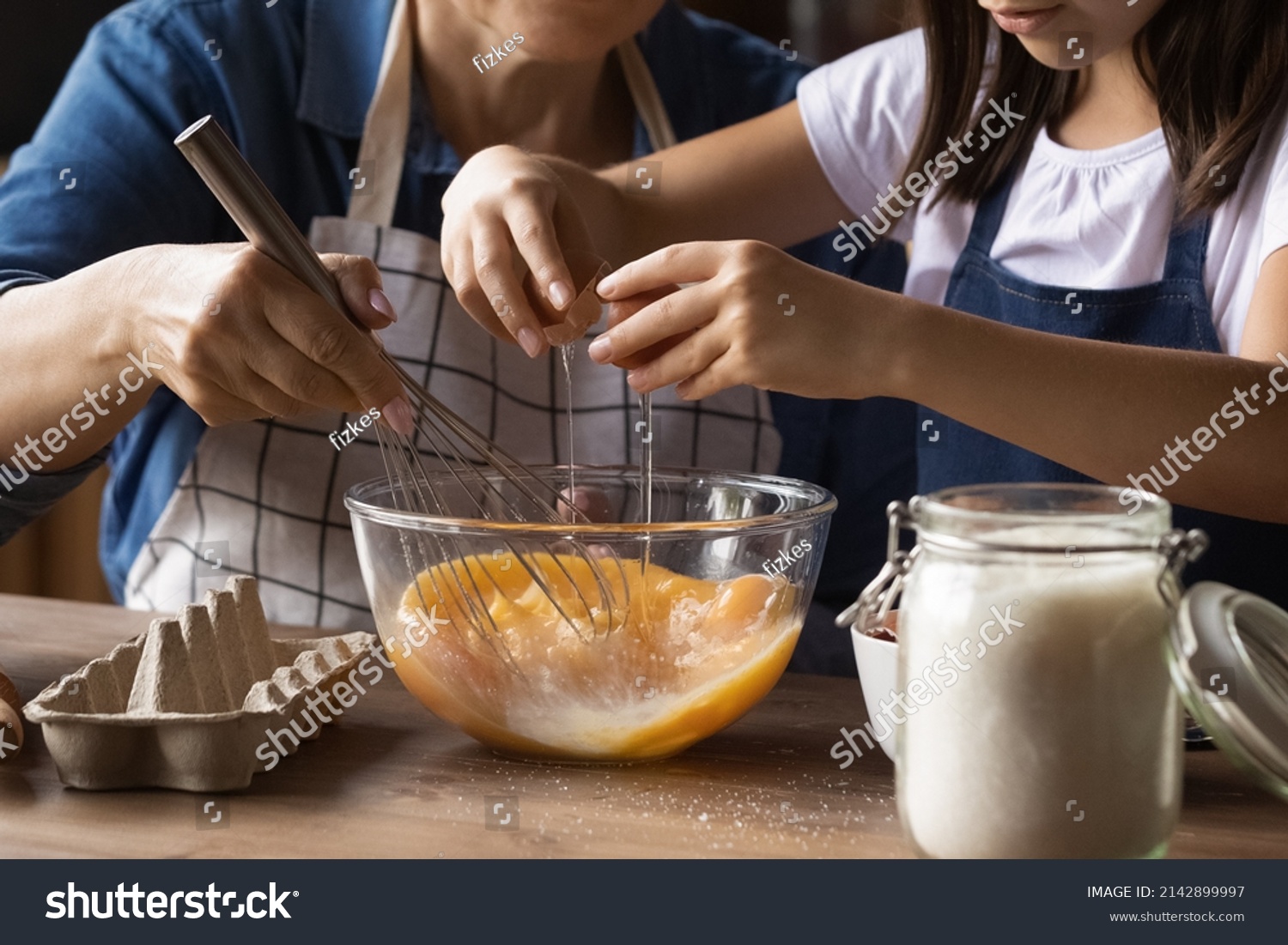 Grandkid helping grandma to cook omelet for breakfast, cracking beating eggs with whisker in bowl at kitchen table. Grandmother teaching grandchild to bake, preparing dough. Family activity. Close up #2142899997