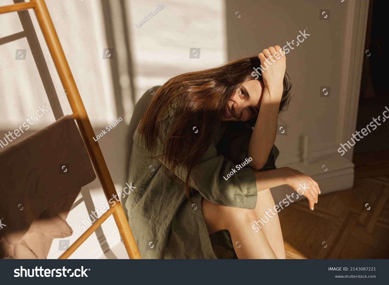 Cute caucasian young woman covers her face with hand from sun while sitting in light room. Brunette in casual home clothes looks at camera. Happy weekend concept #2143087221
