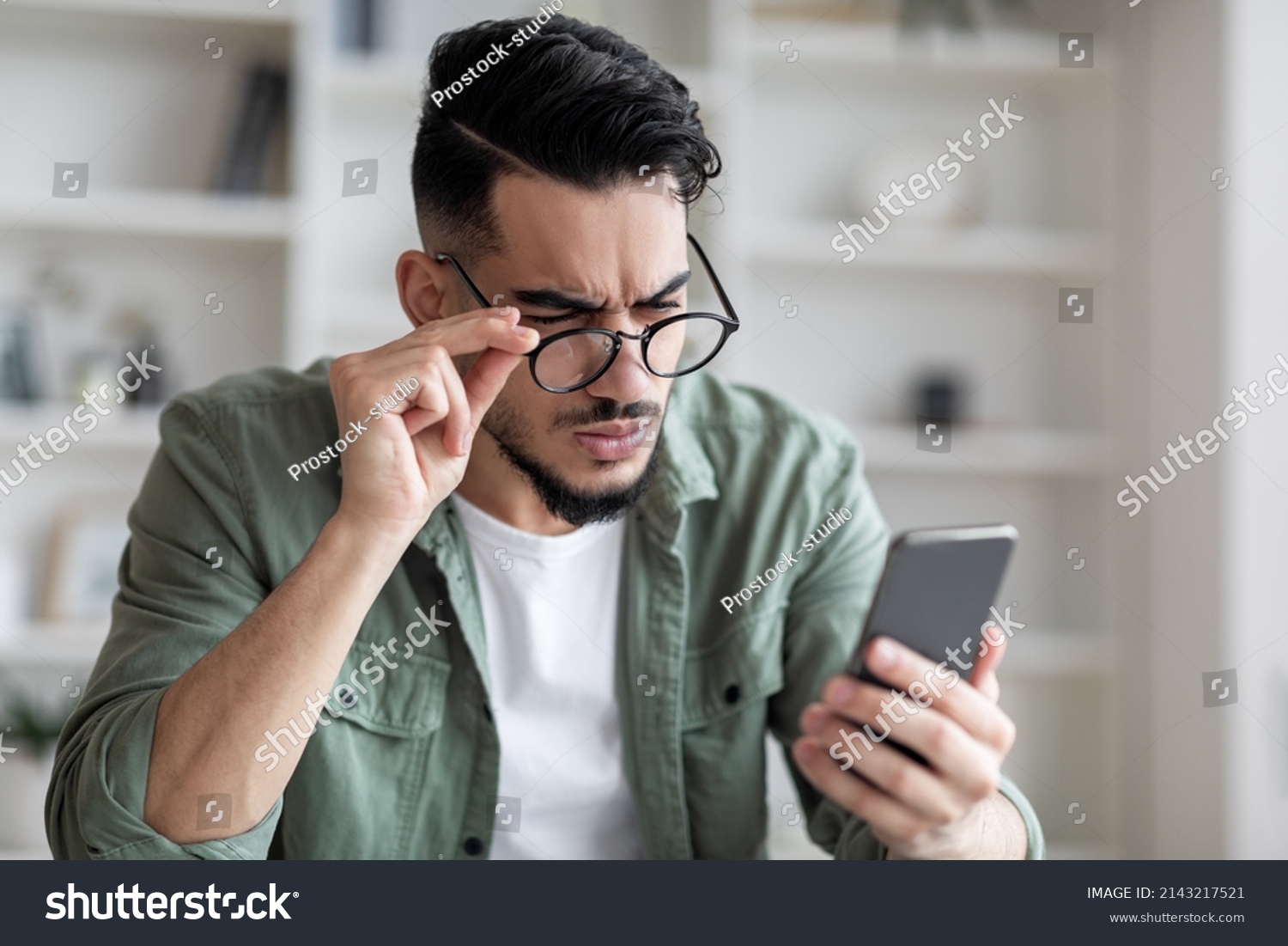 Eyesight Problems Concept. Young Arab Man In Eyeglasses Looking At Smartphone Screen And Frowning, Millennial Guy Trying To Read Message, Suffering From Astigmatism And Bad Vision, Closeup Shot #2143217521