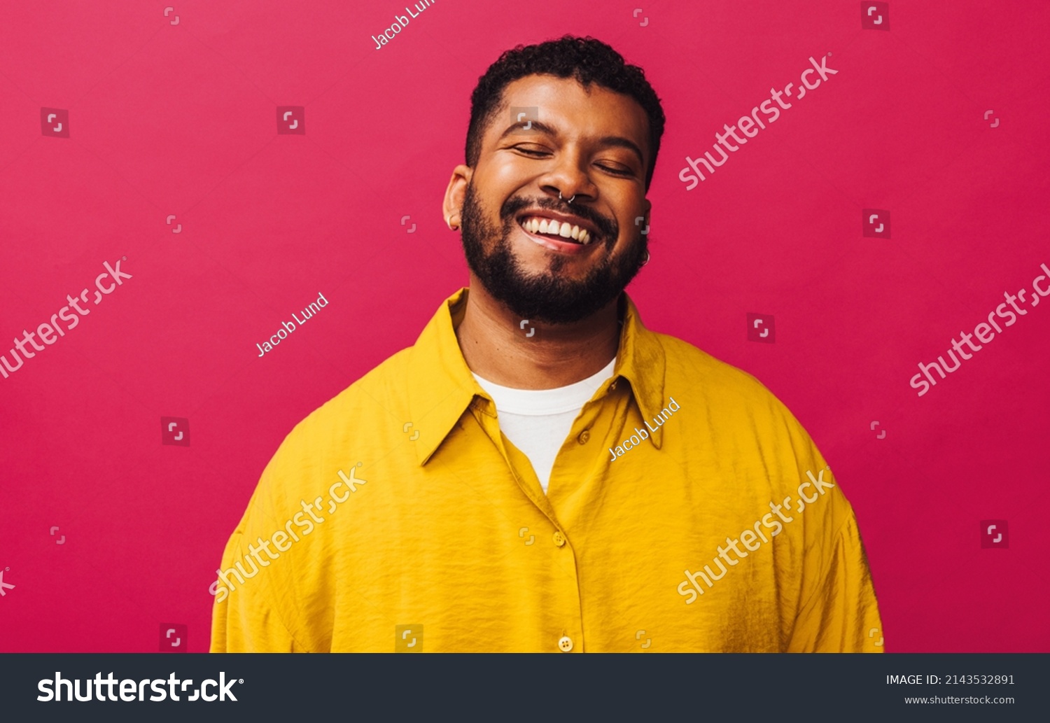 Happy young man smiling cheerfully with his eyes closed in a studio. Handsome young man with piercings standing alone against a pink background. Stylish young man feeling vibrant. #2143532891