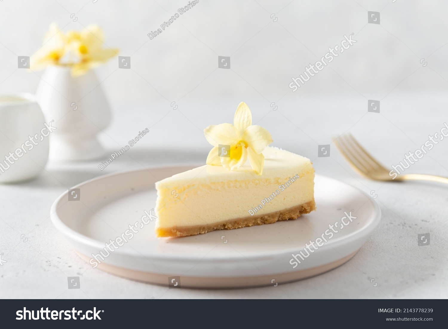 Classic New York cheesecake with fresh vanilla flower on a white concrete background, side view. A piece of Vanilla cheesecake on a white plate. Confectionery menu, recipe. Close up. #2143778239