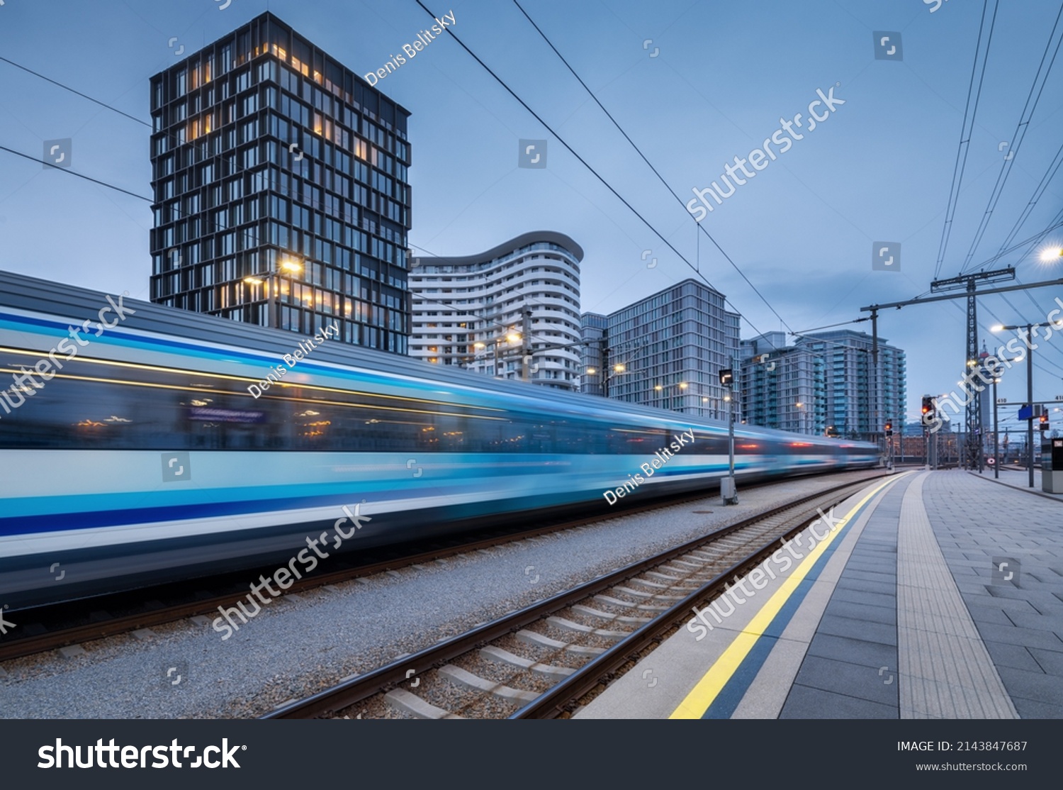 High speed train in motion on the railway station at dusk. Moving blue modern intercity passenger train, railway platform, buildings, city lights. Railroad in Vienna, Austria. Railway transportation #2143847687