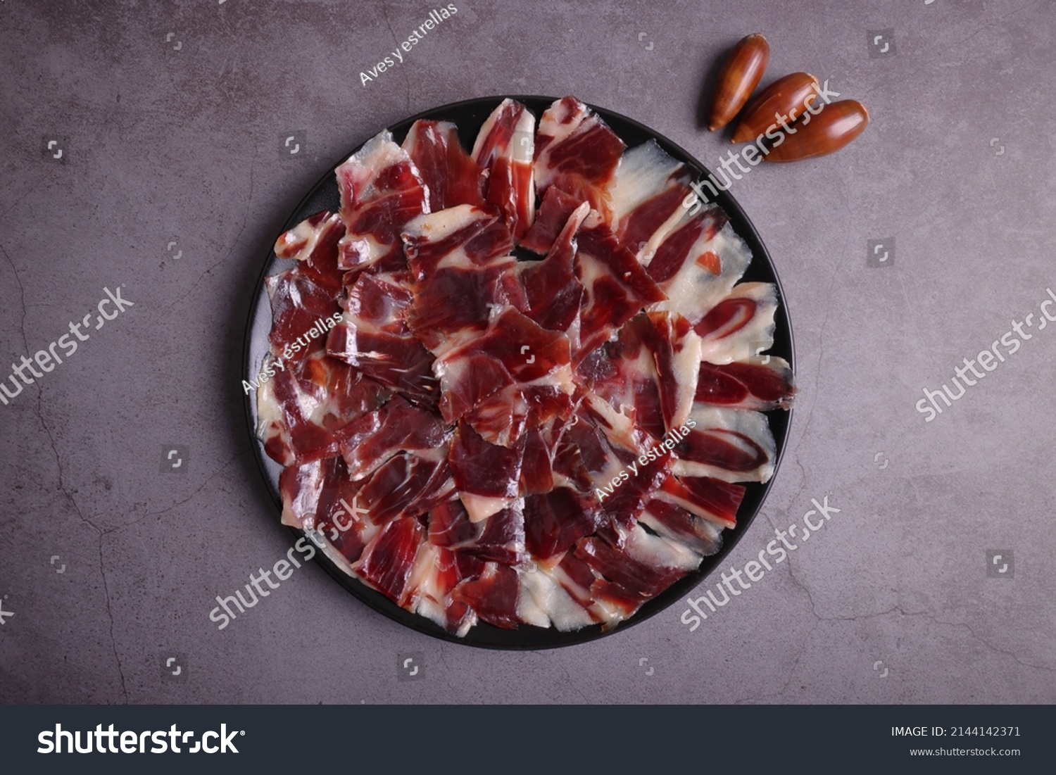 Portion of acorn-fed Iberian ham 100% Dehesa de Extremadura on a black plate, on a dark gray table and decorated with acorns #2144142371