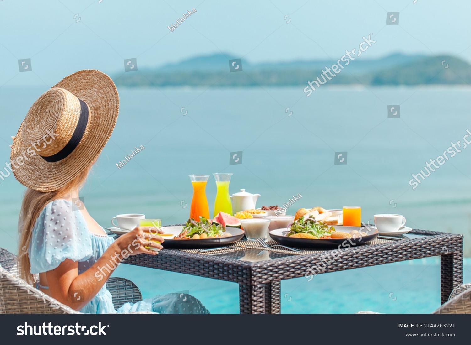 Dinner with sea view in luxury hotel. Woman in straw hat near swimming pool, eating food and enjoy ocean view. Dinner table on tropical vacation. Back view. Concept of travel, holidays, weekend. #2144263221