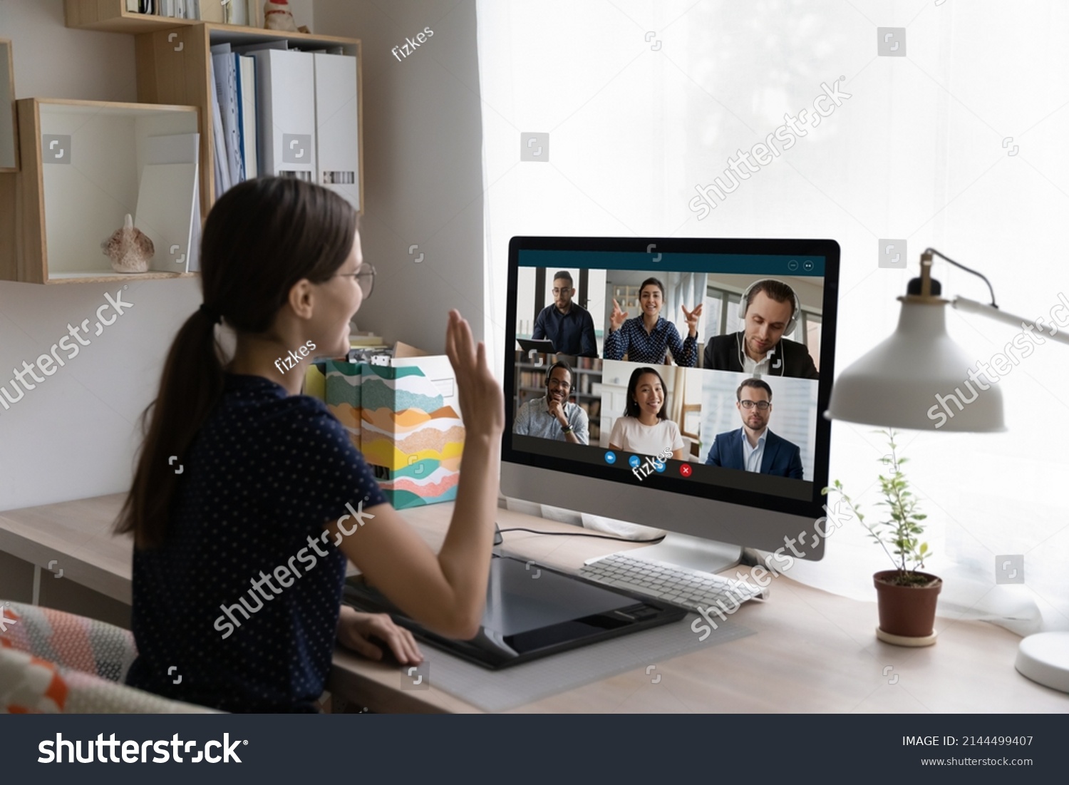 Young woman sit at desk in front of pc, looks at screen, wave hand greeting diverse friends, multinational colleagues start or finish group video call. Virtual meeting, remote communication concept #2144499407