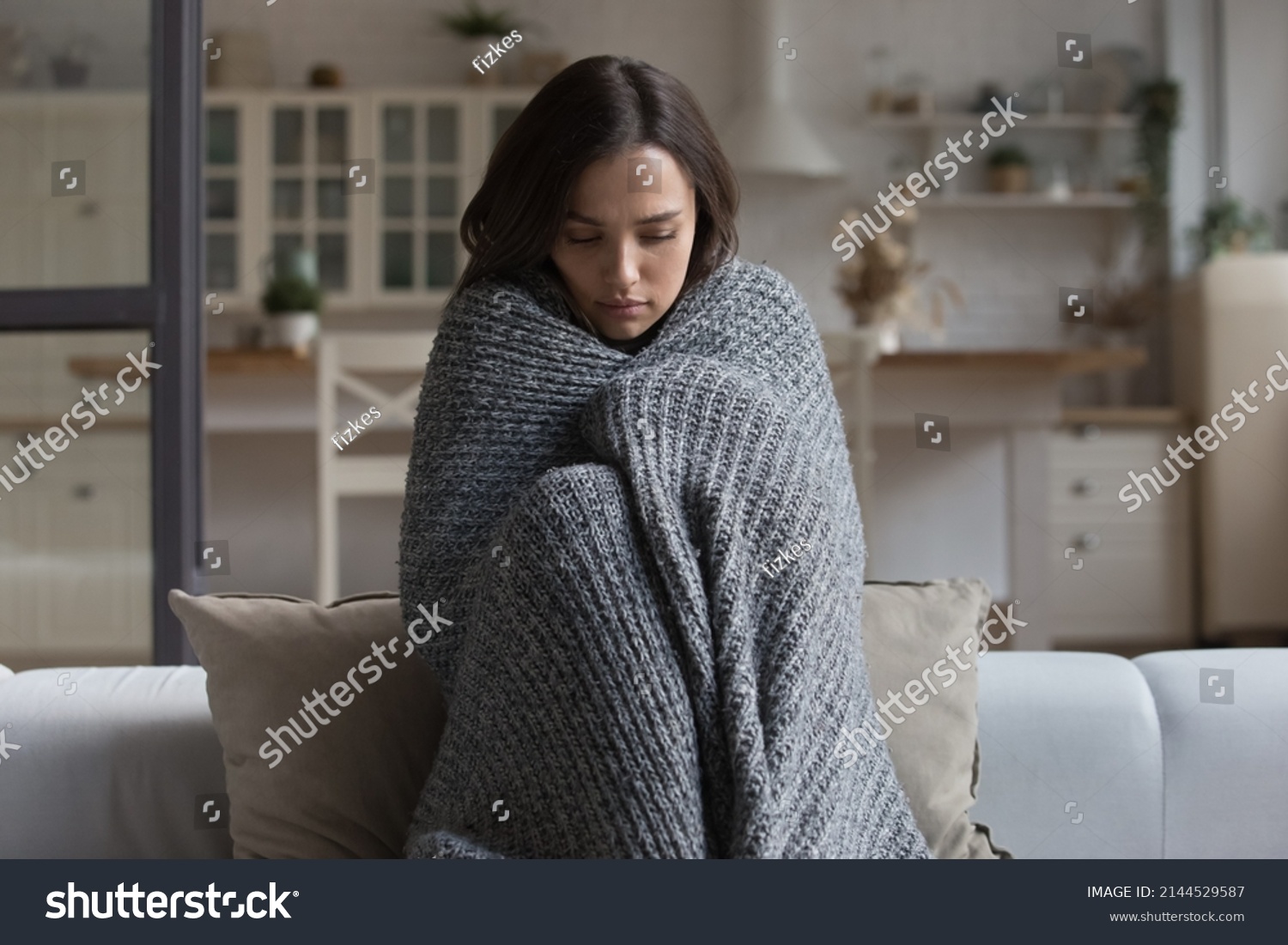 Stressed sad wrapped in plaid young woman feeling cold, ill, sick, suffering from fever, virus, trying to warm, sitting on couch at home. Frustrated shocked girl going through depression #2144529587