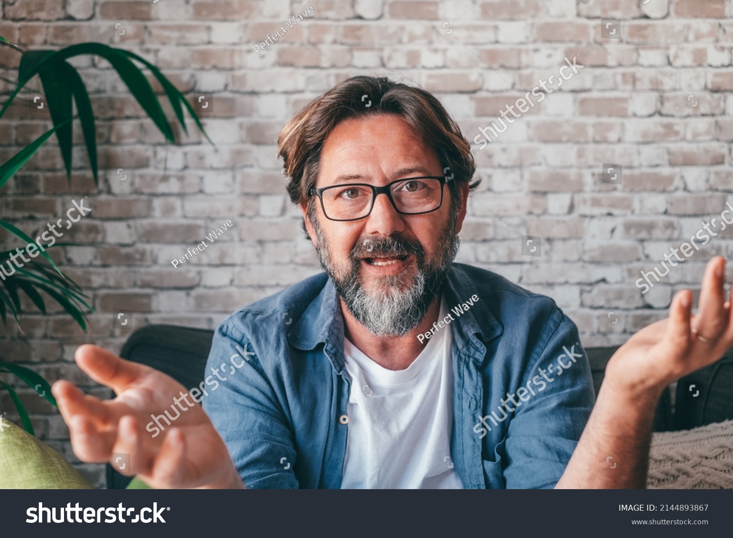 Bearded middle aged man in casual clothes and glasses gesticulating and talking to camera while sitting on sofa against brick wall in daytime at home #2144893867