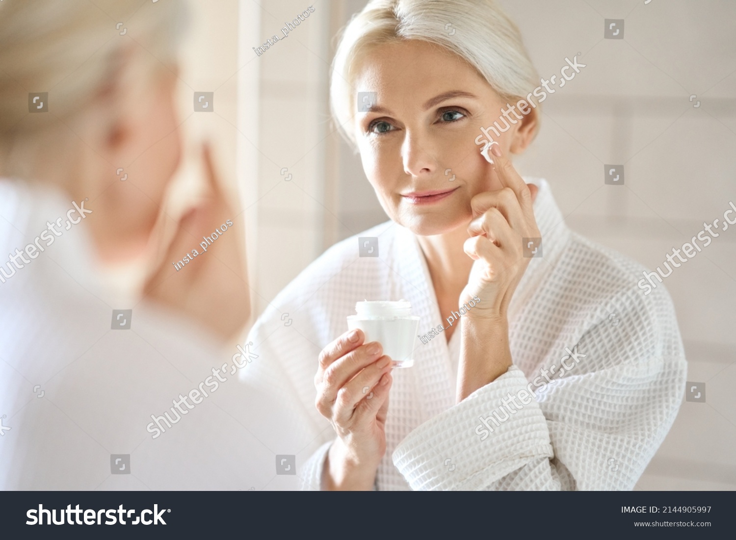 Attractive mid age older adult 50 years old blonde woman wears bathrobe in bathroom applying nourishing antiage face skin care cream treatment, looking at mirror doing daily morning beauty routine. #2144905997