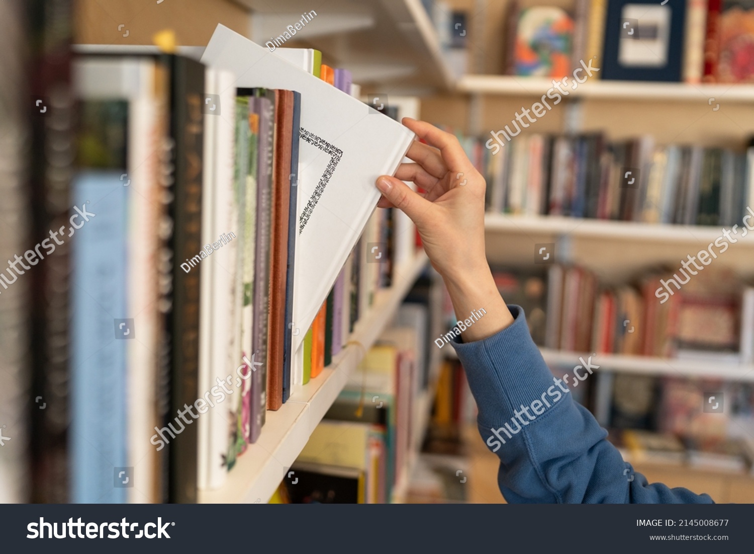 Female hand pulling book from bookshelf in public library in university, college or high school. Woman student take novel from bookcase in bookshop store, soft focus. Education and literature concept #2145008677
