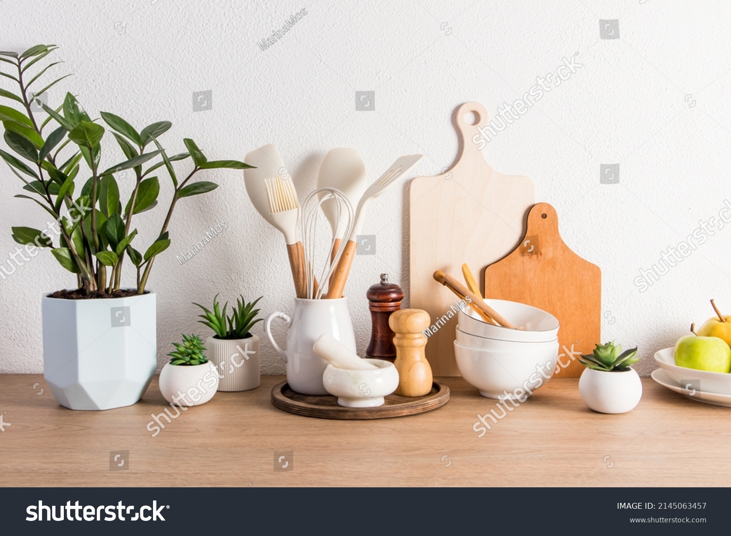 a variety of kitchen utensils, utensils, a potted flower and fruit on a plate on a wooden countertop against a white textured wall. front view #2145063457