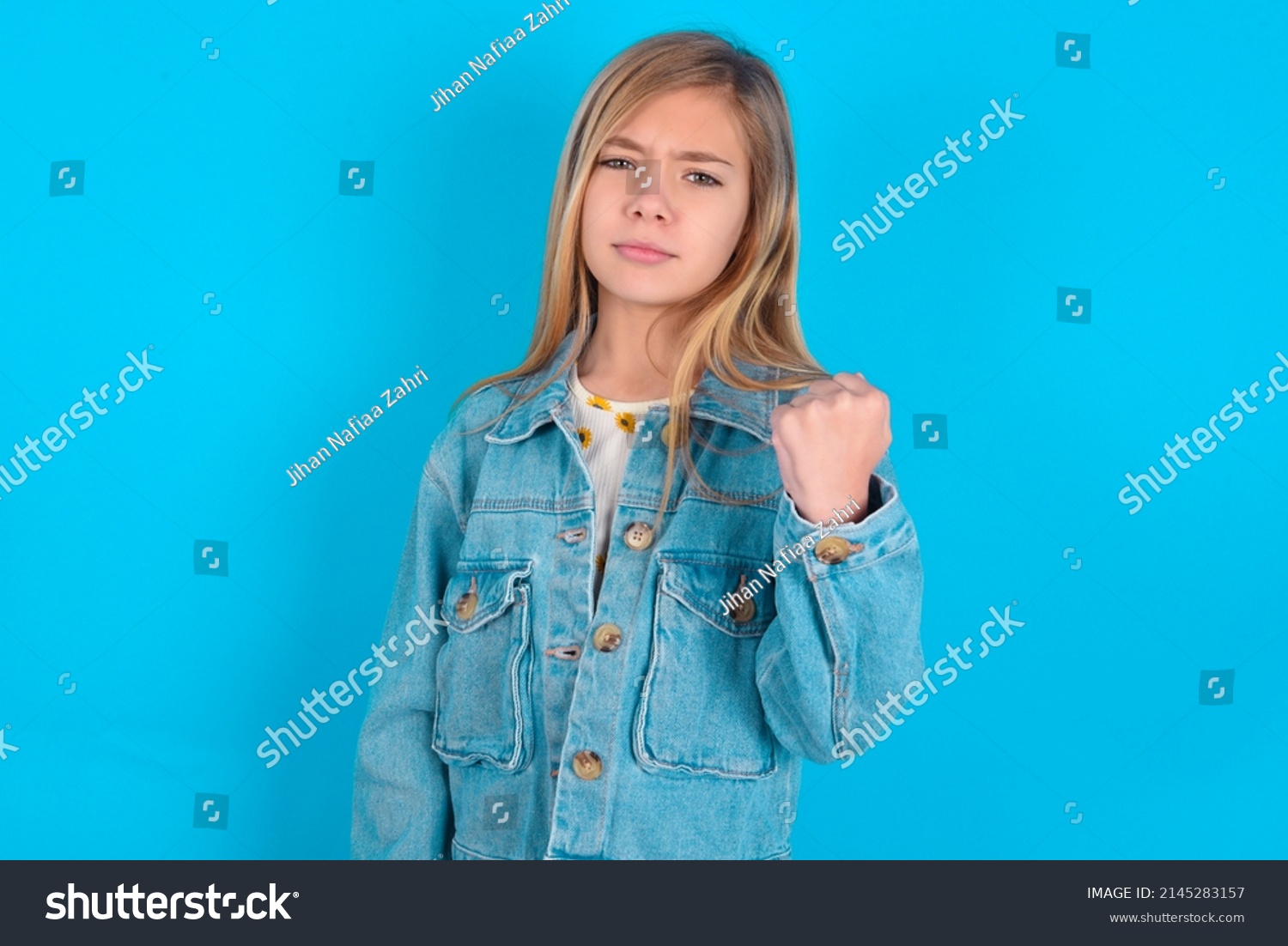 blonde little kid girl wearing denim jacket over blue background shows fist has annoyed face expression going to revenge or threaten someone makes serious look. I will show you who is boss #2145283157