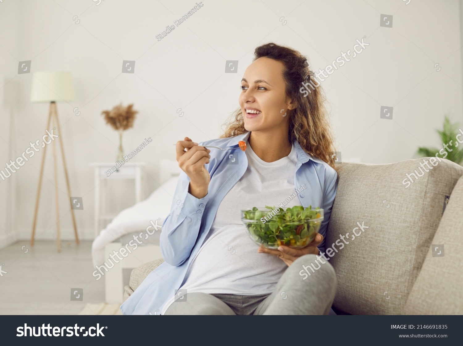 Pregnant woman who eats many different vegetables during pregnancy enjoys fresh green salad at home. Happy woman eating salad for breakfast sitting on sofa in living room. Concept of healthy pregnancy #2146691835