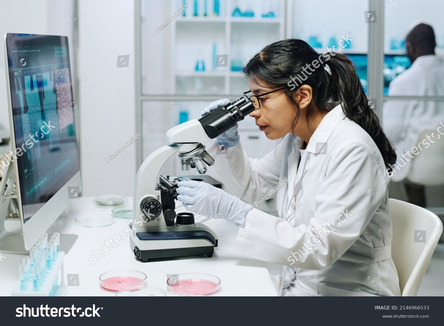 Side view of female biochemist in whitecoat, gloves and eyeglasses looking in microscope while studying virus in laboratory #2146966533