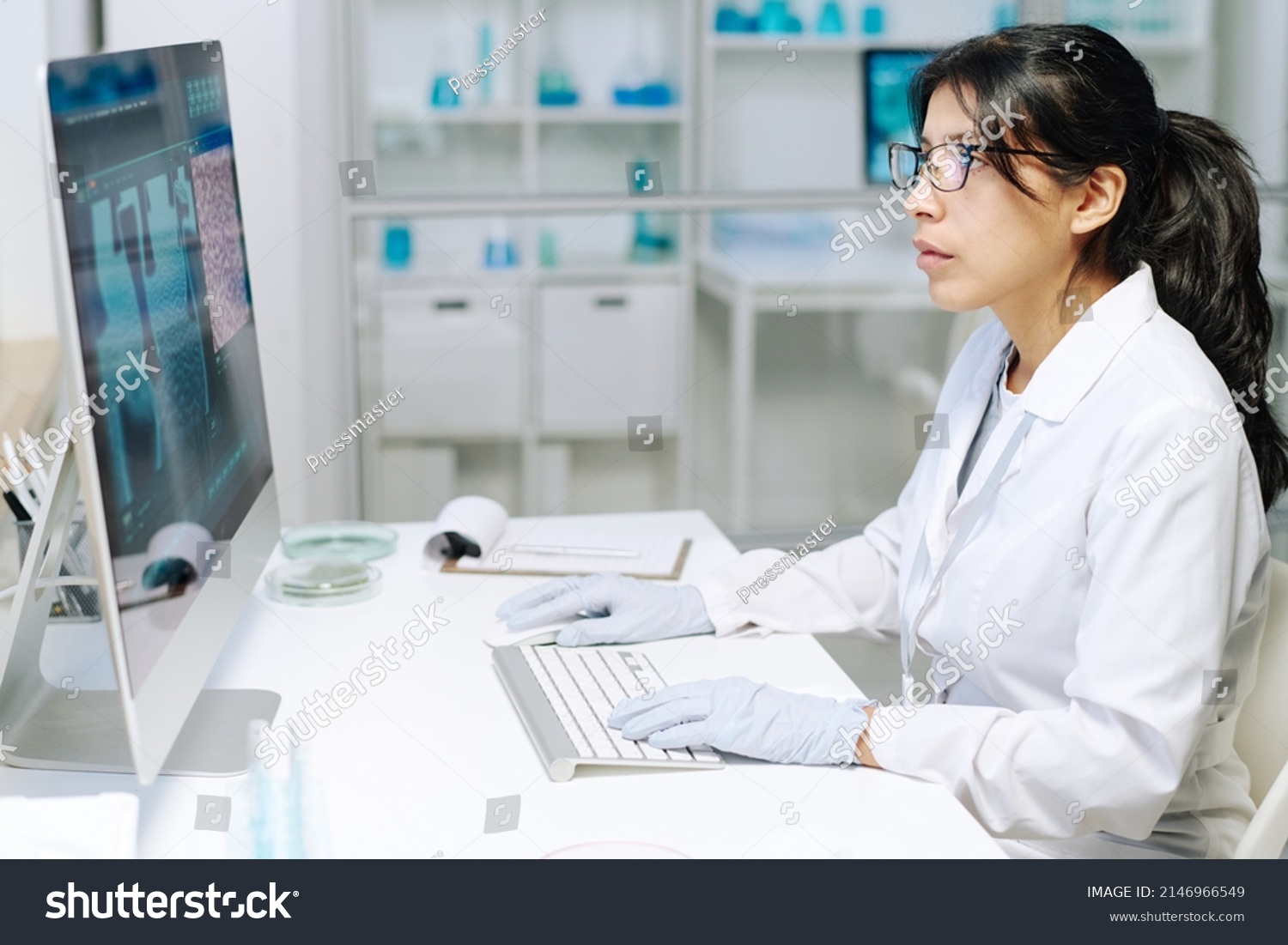 Young serious female researcher or virologist in whitecoat and gloves sitting in front of computer screen while analyzing new strain #2146966549