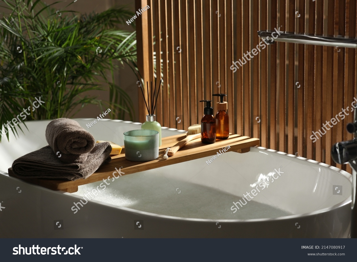 Wooden bath tray with candle, air freshener and bathroom amenities on tub indoors #2147080917