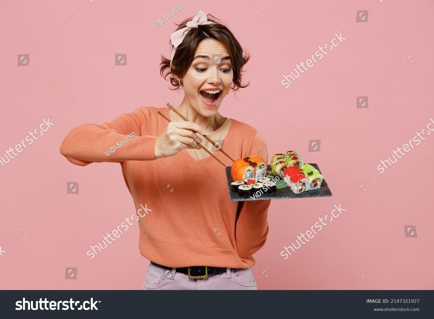 Young excited amazed cool fun woman 20s in casual clothes hold in hand eat makizushi sushi roll served on black plate traditional japanese food chopsticks isolated on plain pastel pink background. #2147321927