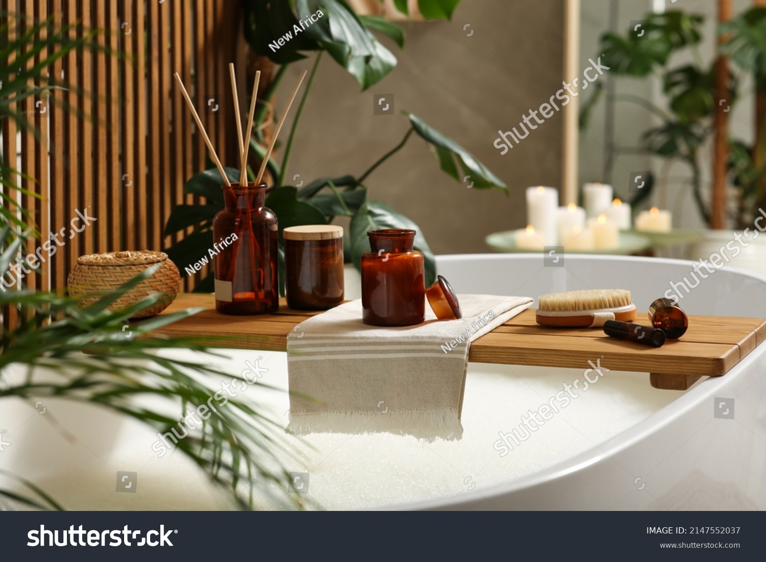 Wooden bath tray with open book, candle and body care products on tub indoors. Relaxing atmosphere #2147552037