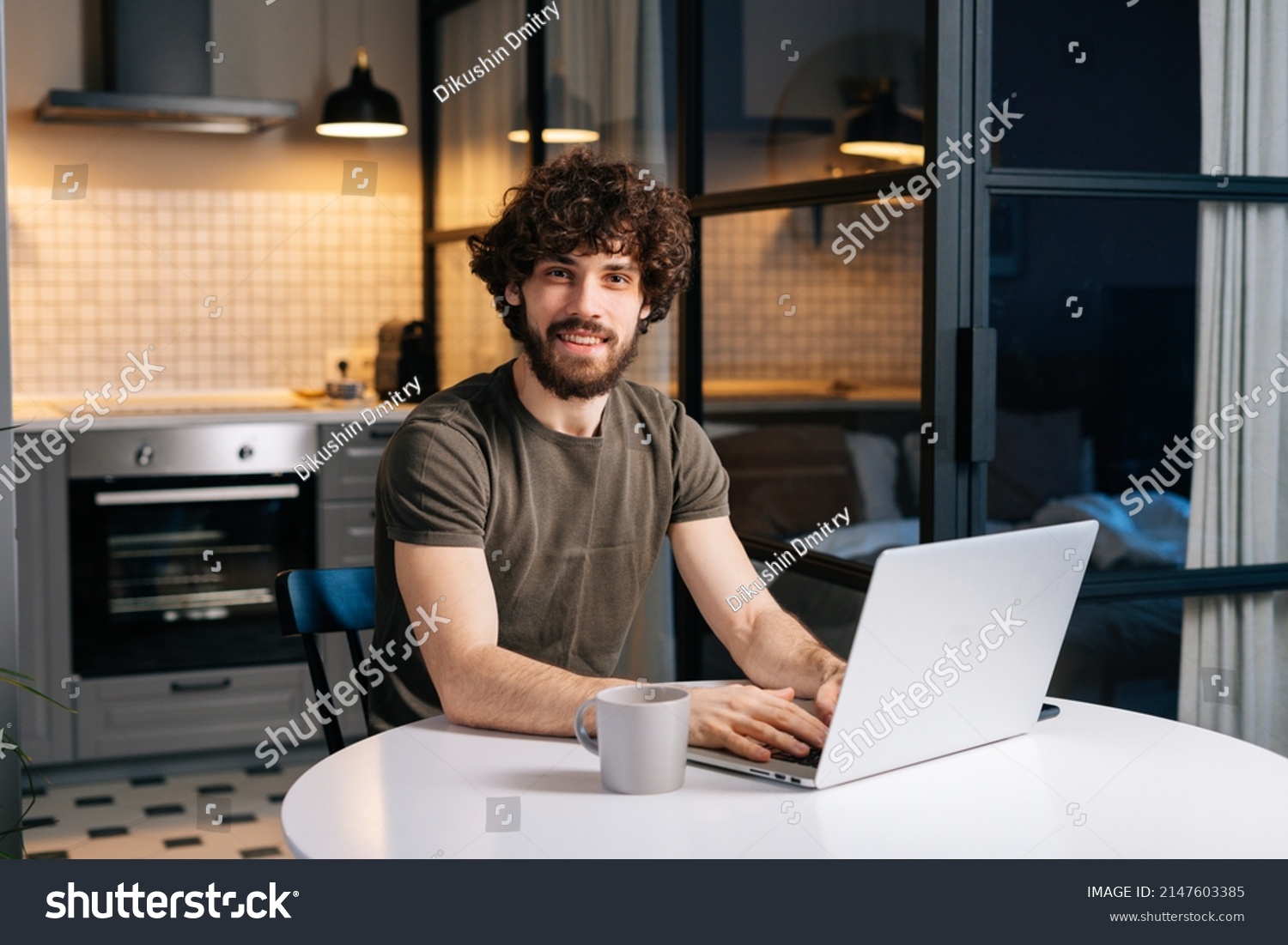 Portrait of cheerful bearded young man using typing on laptop sitting at table in kitchen room with modern interior, looking at camera. Confedent curly happy freelancer male remote working from home. #2147603385