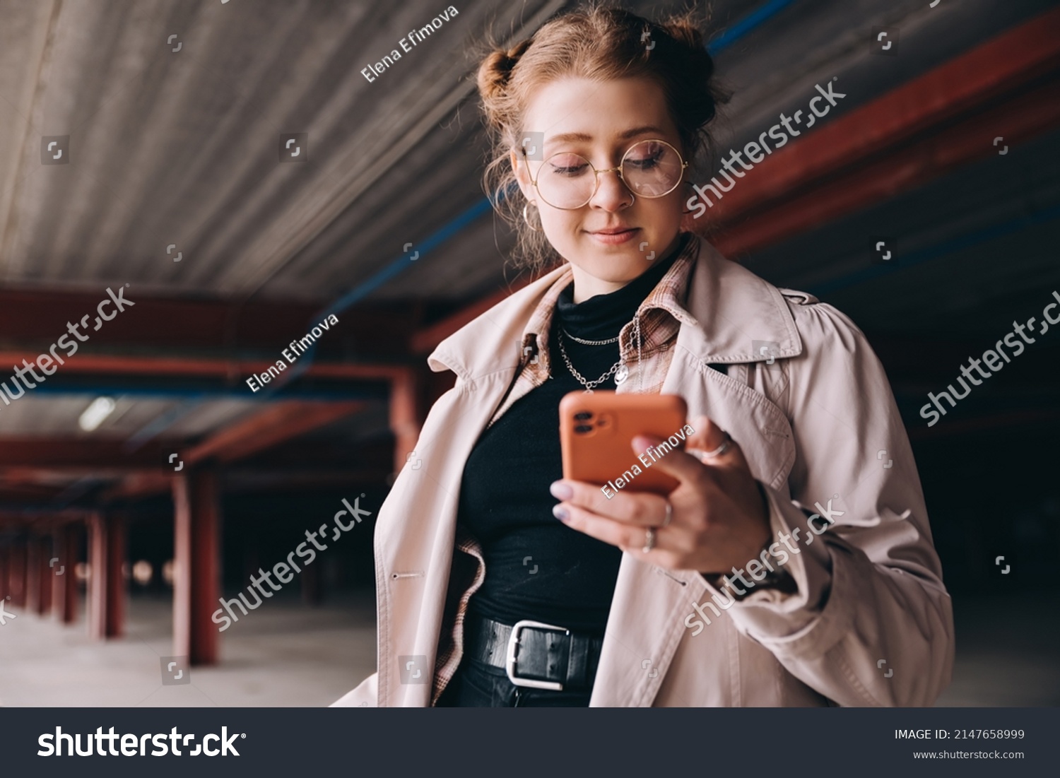 young caucasian female student, generation z, dressed in a trench coat in the parking lot uses a smartphone #2147658999