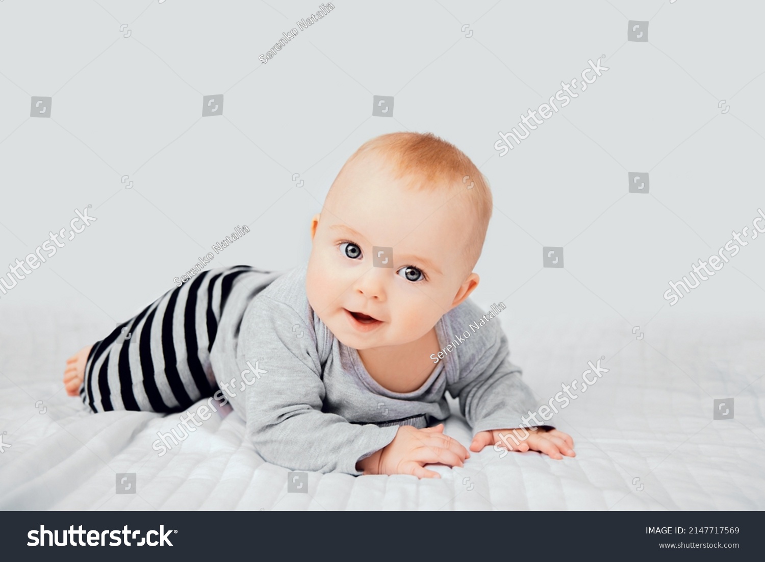 Seven month old baby child sitting on bed. Cute smiling little infant girl on white soft blanket. Charming blue eyed baby. Copy space. #2147717569