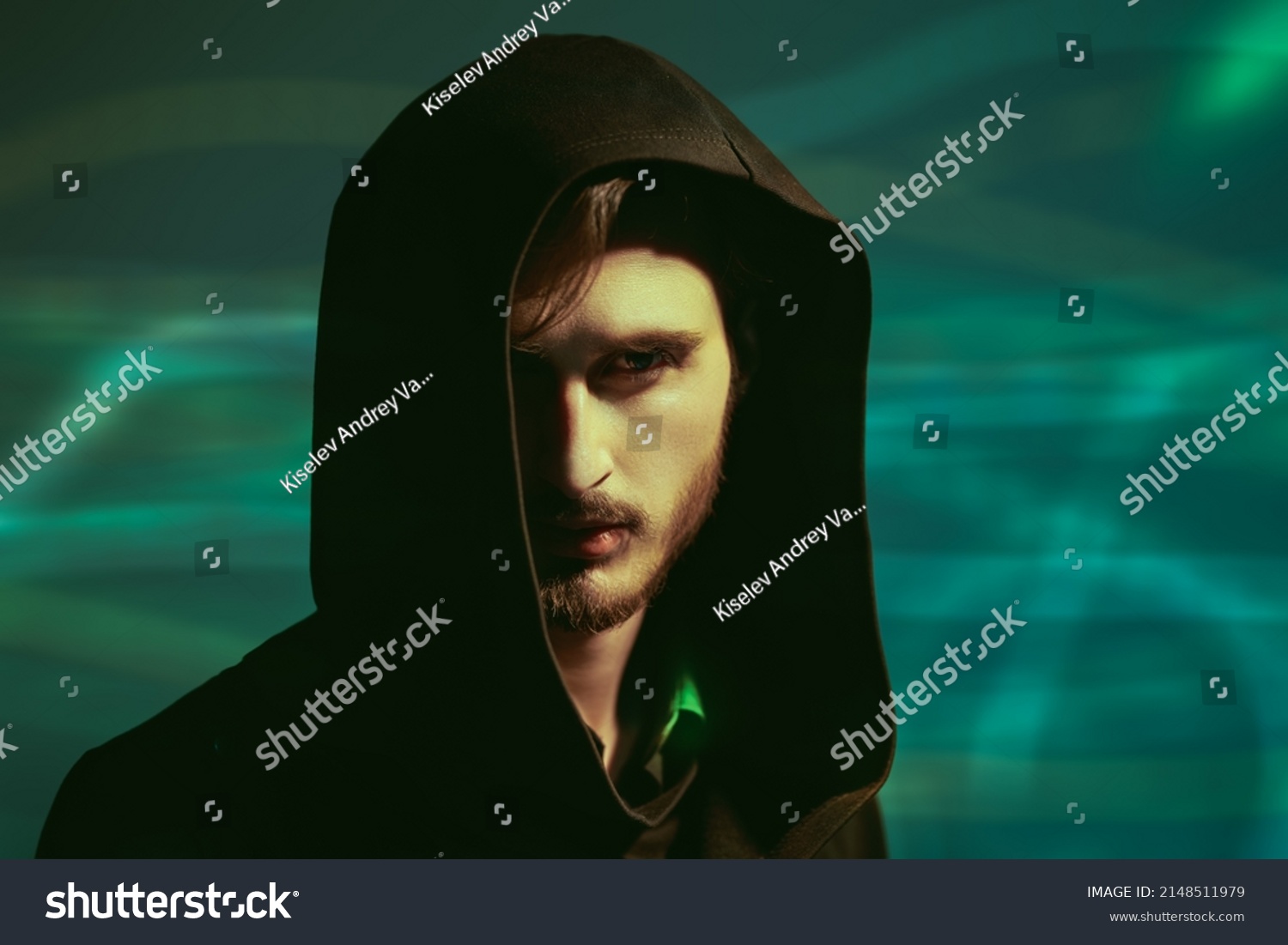 Studio portrait of a brutal brunette man looking intently and cautiously at the camera from under a black hood. Green background. People and emotions.  #2148511979