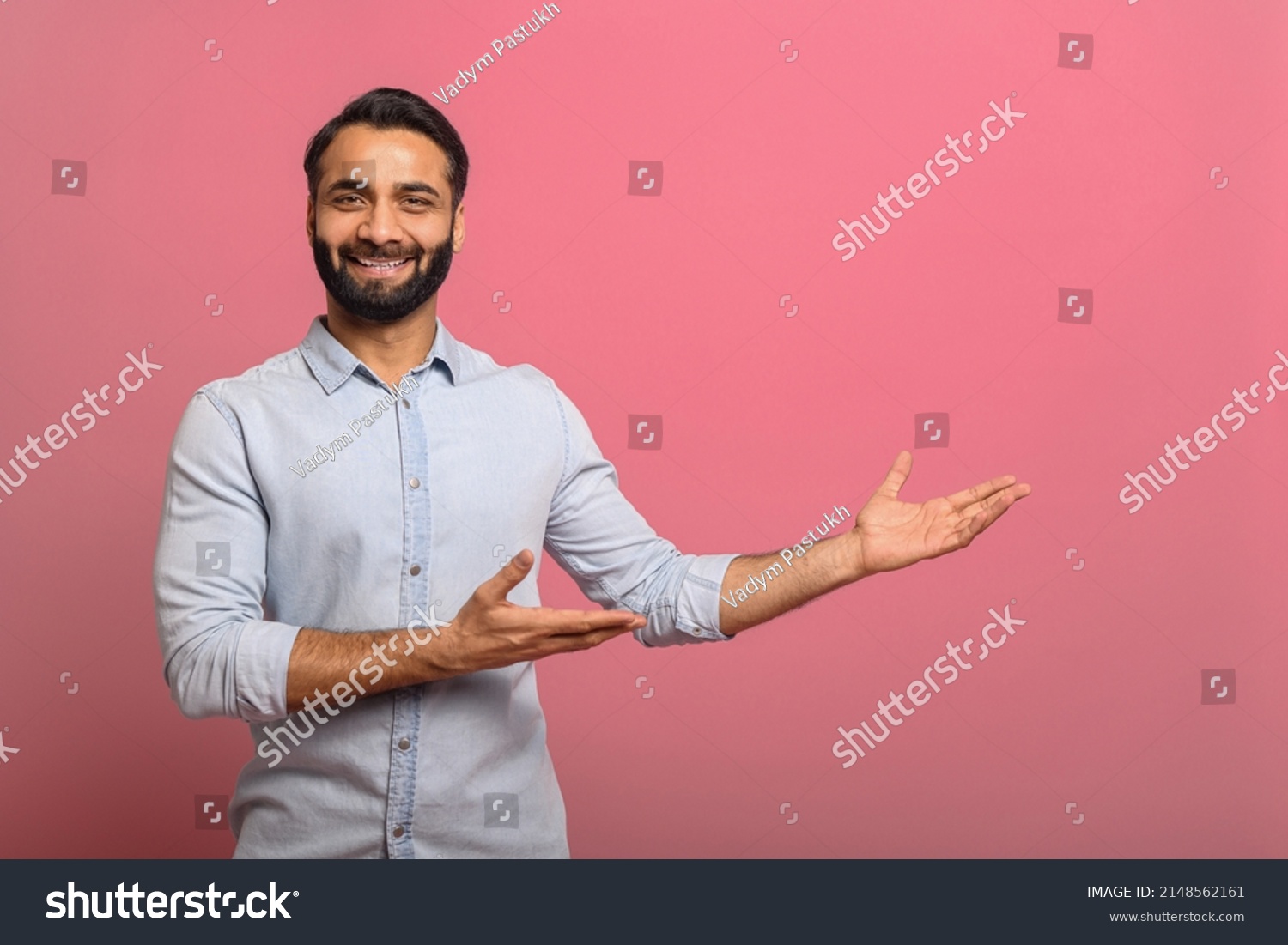 Handsome Indian guy points palms at empty copy space isolated on pink background. Cheerful multiracial bearded man in casual jeans shirt presenting novelty, advertising concept #2148562161
