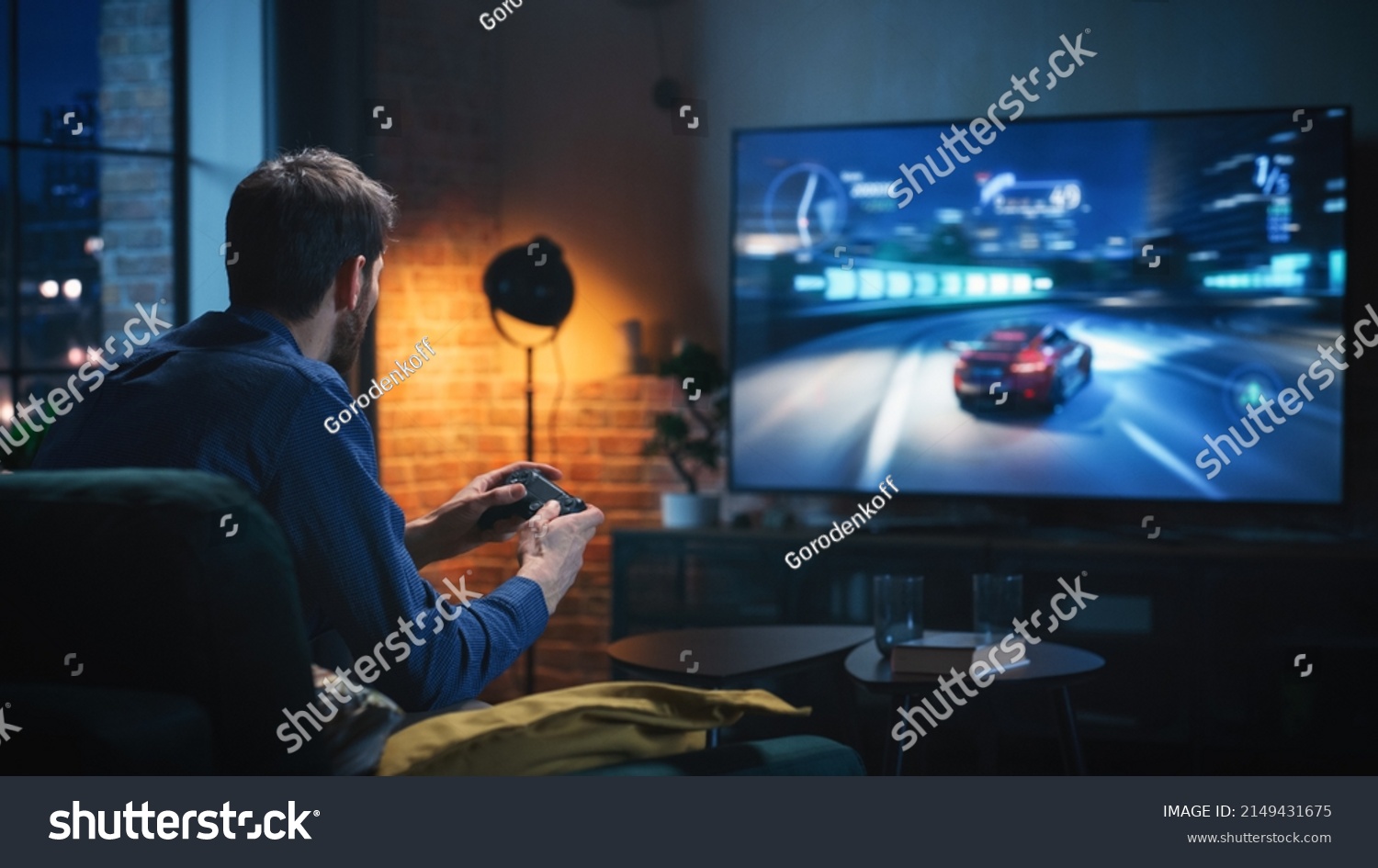 Young Man Spending Time at Home, Sitting on a Couch in Stylish Loft Apartment and Playing Arcade Car Video Games on Console. Male Using Controller to Play Street Racing Drift Simulator. #2149431675