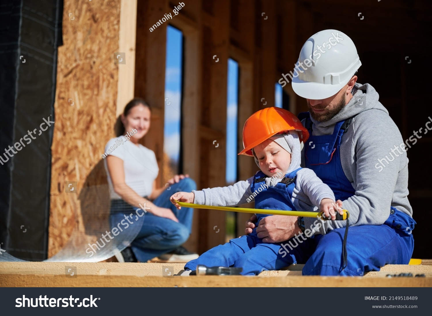 Father, mother and son building wooden frame house. Toddler boy helping his daddy, while woman looking for them on construction site. Guys wearing helmet and blue overalls. Carpentry, family concept. #2149518489