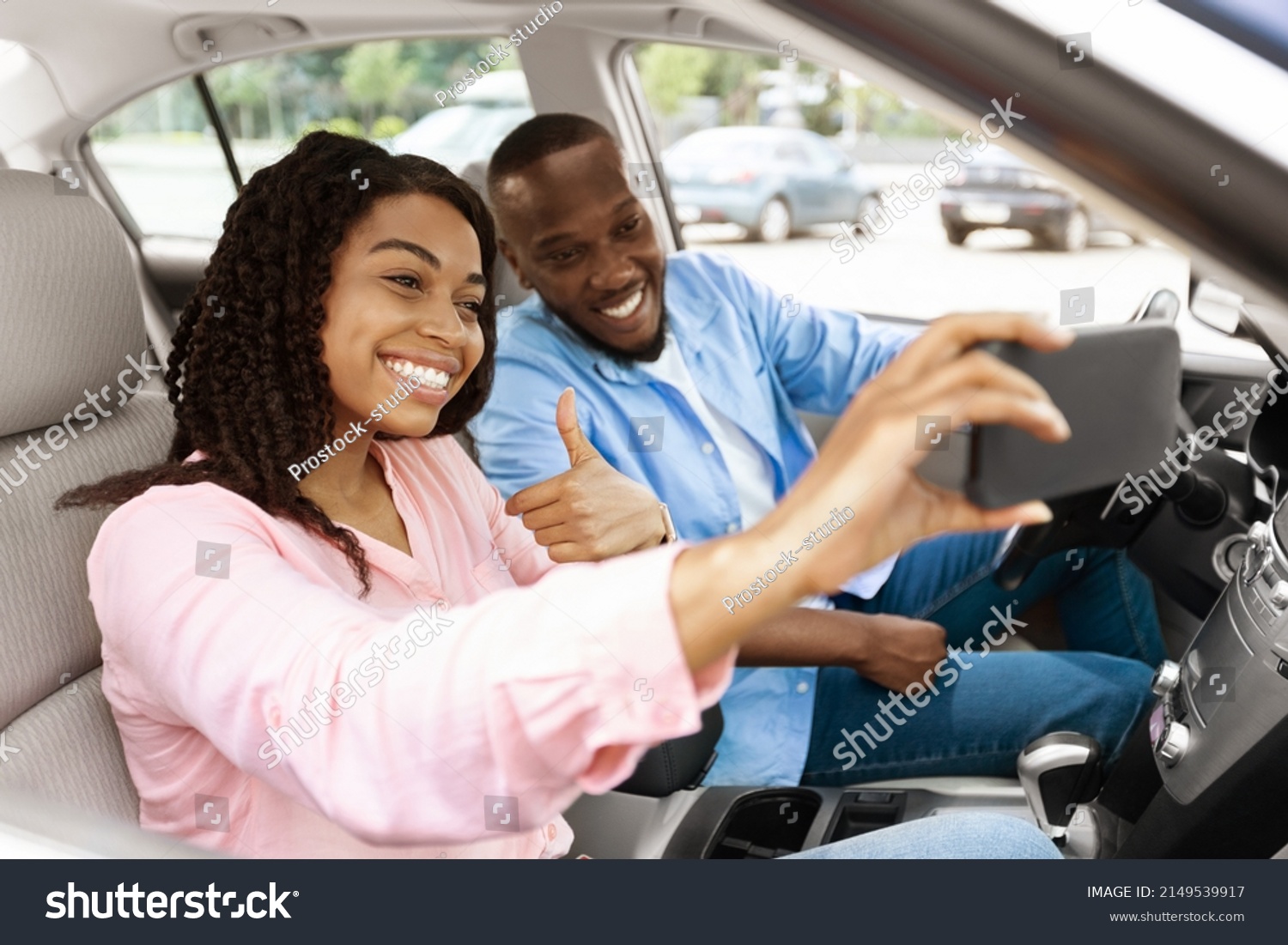 Beautiful young African American couple taking selfie inside car, using mobile phone, showing thumbs up sign gesture. Happy black man and woman going on vacation, taking self photo, buying new auto #2149539917