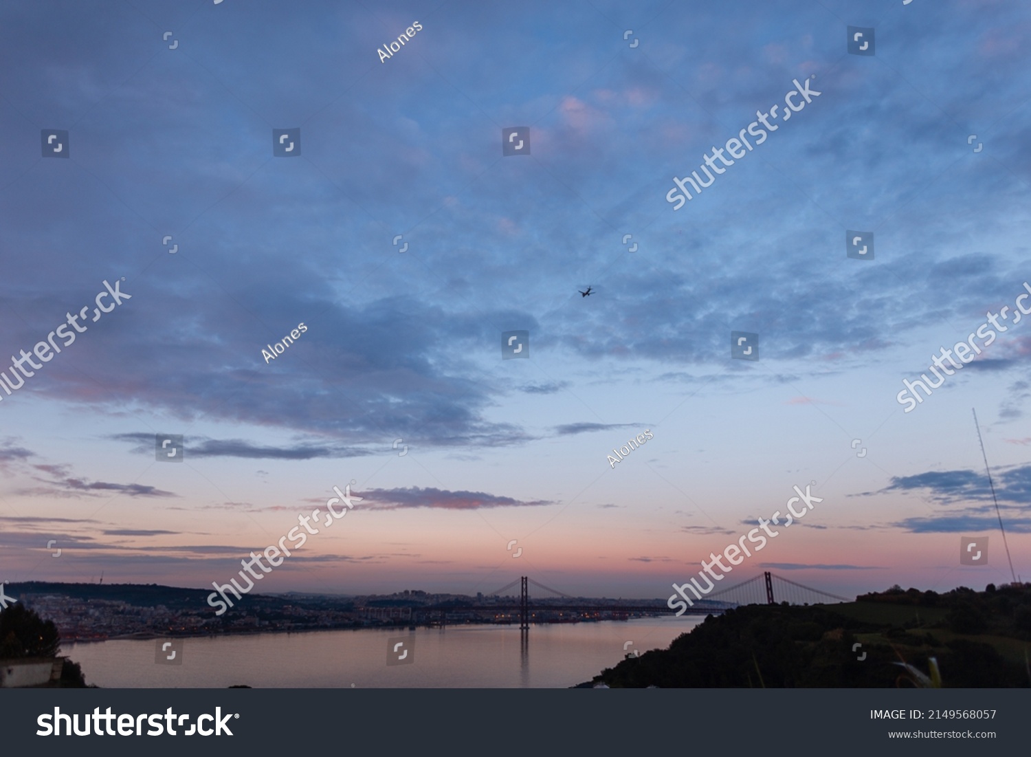 Beautiful European Lisbon, Portugal after sunset with evening sky and purple blue clouds. Evening cityscape with bridge, river and airplane  #2149568057