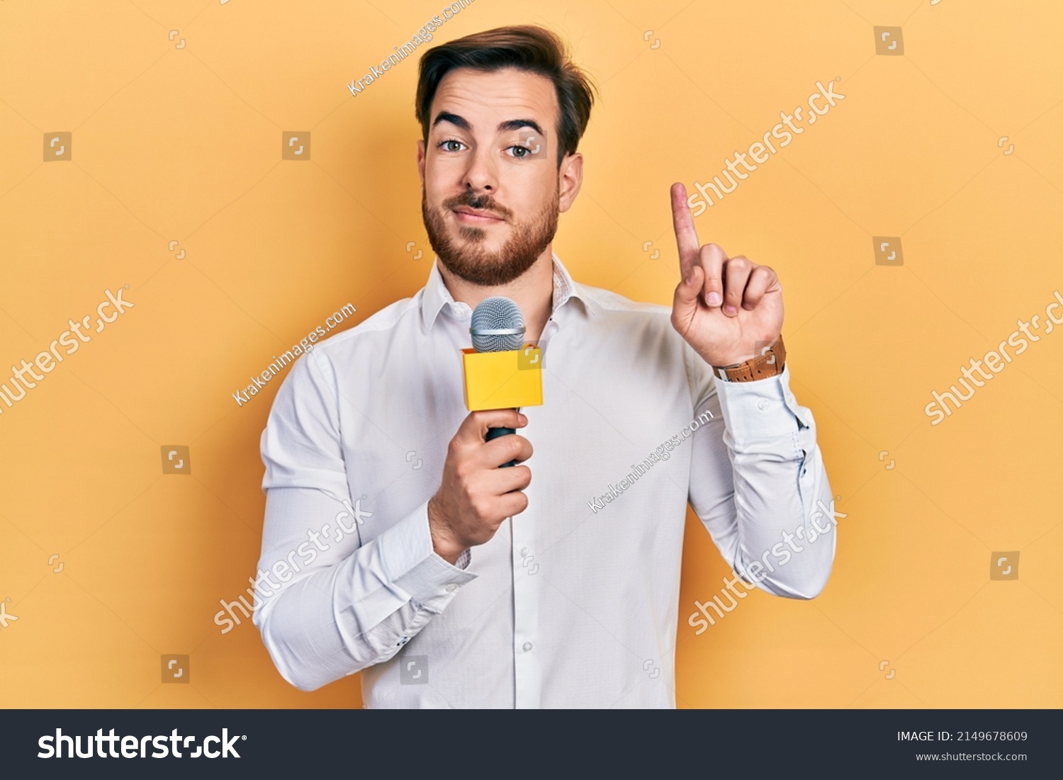 Handsome caucasian man with beard holding reporter microphone surprised with an idea or question pointing finger with happy face, number one  #2149678609