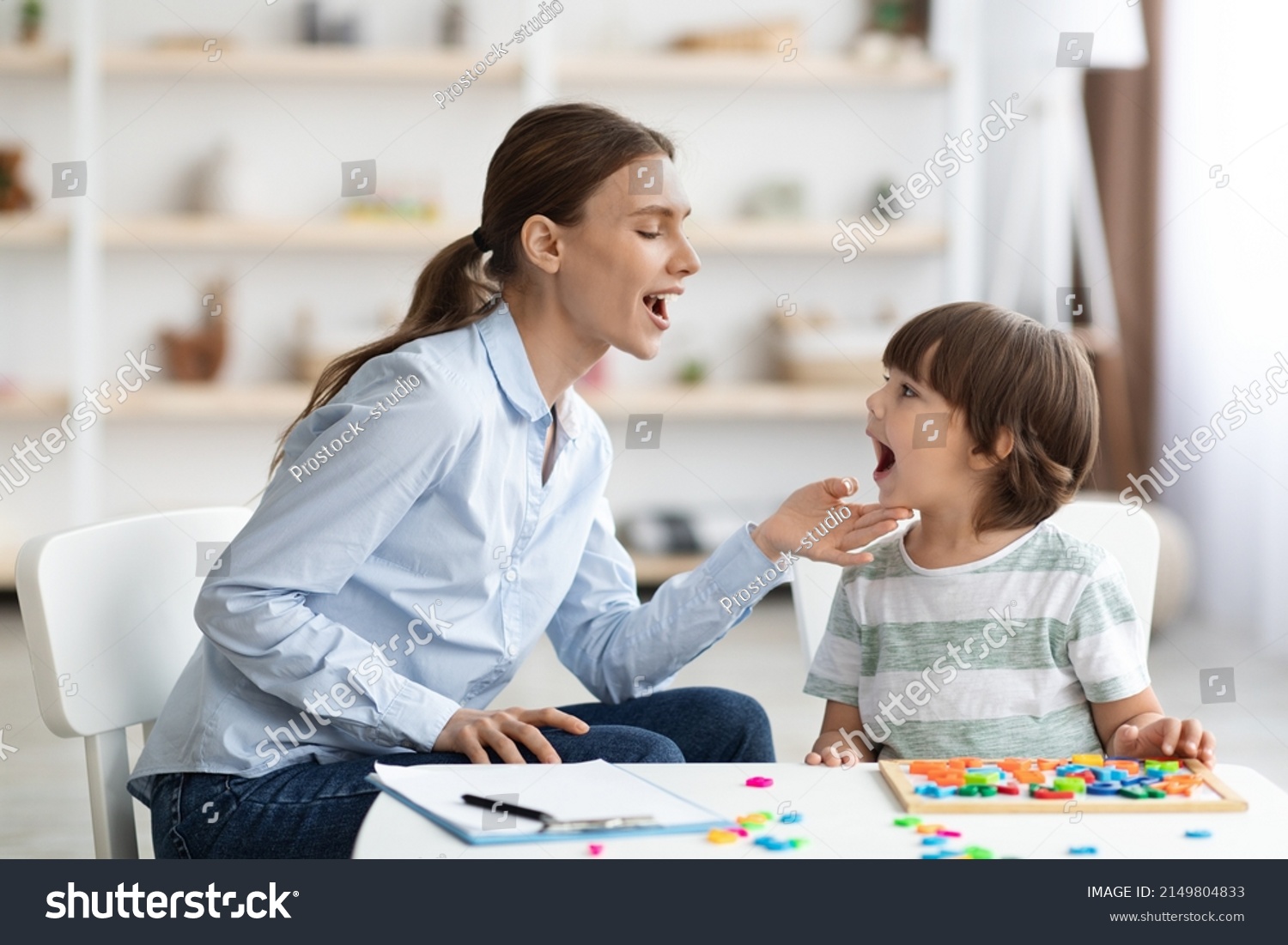 Professional woman speech therapist helping little boy to pronounce right sounds, showing mouth articulation at office, free space #2149804833