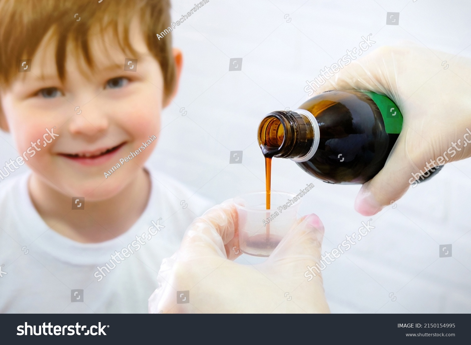 Happy little boy, smiling, wants to take the cure for the disease. Hands pouring cough medicine into a special measuring cap. Horizontal photo. #2150154995