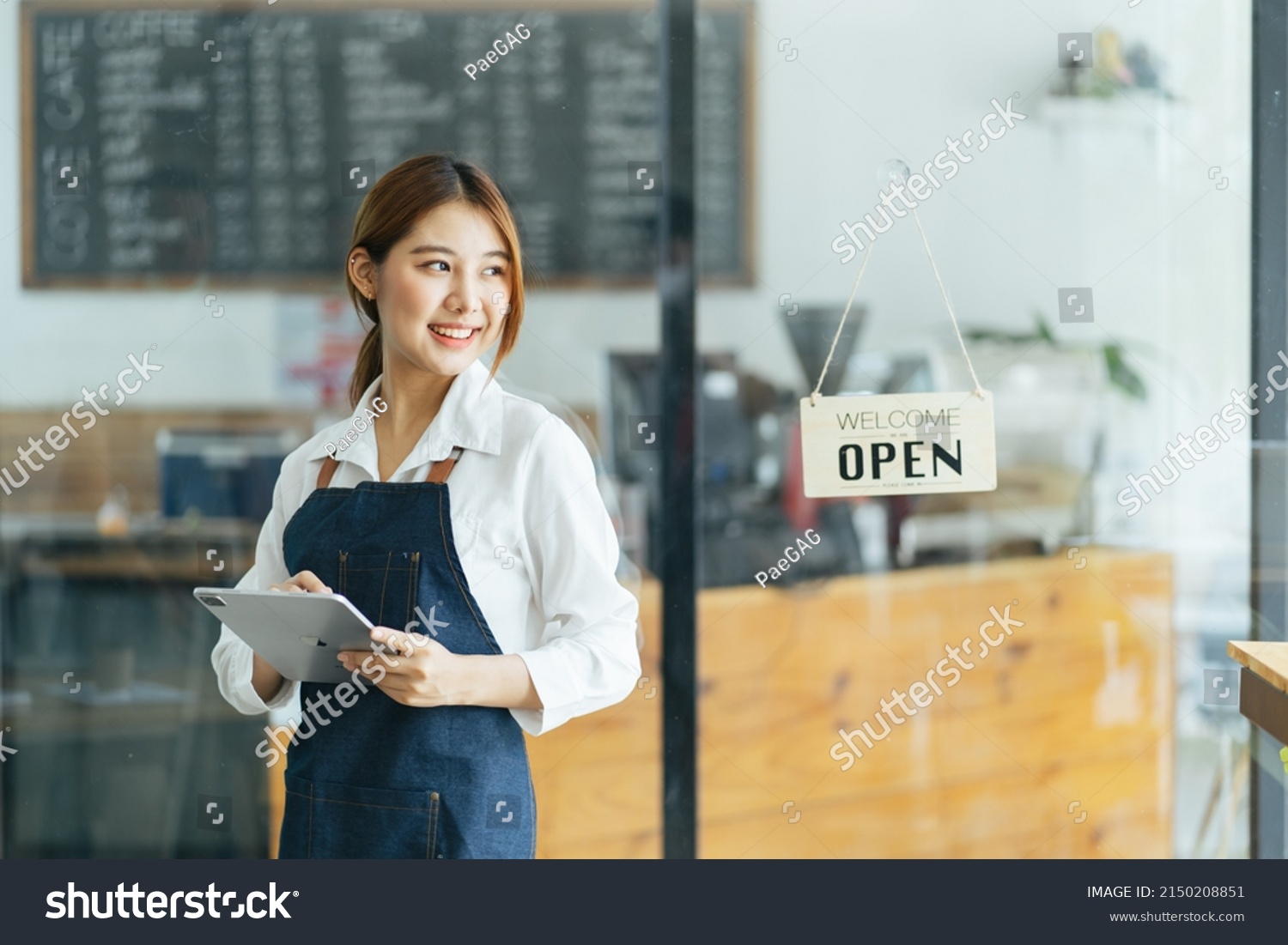 Portrait of happy woman standing at doorway of her store. Cheerful mature waitress waiting for clients at coffee shop. Successful small business owner in casual wearing blue apron standing at entrance #2150208851