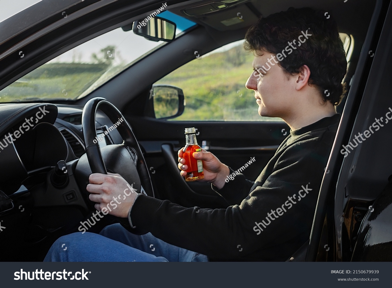 The driver, a young man driving a car, drinks an alcoholic brandy drink from a bottle  #2150679939