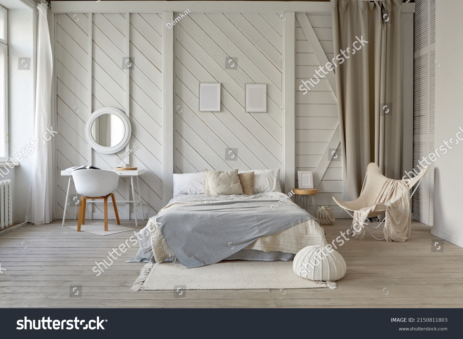 Comfortable bed with blanket and cushions placed on rug at wall with decorative frames in stylish bedroom with table and armchair in pastel colors #2150811803