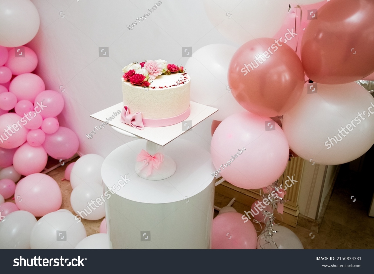 An appetizing festive yellow cake adorned with vibrant red, white and pink flowers sits on a white decorative stand in a room adorned with pink and white balloons. #2150834331