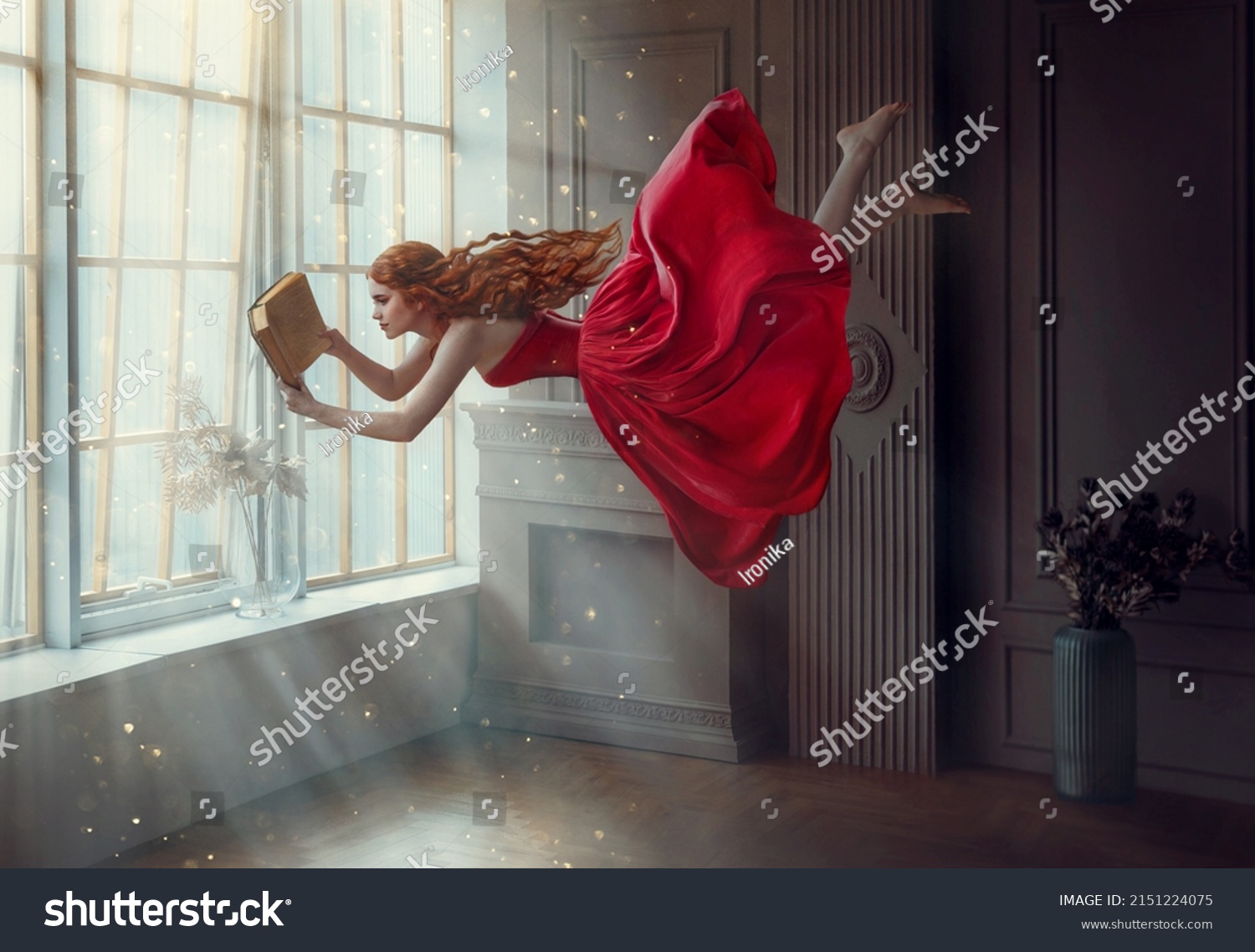 Fantasy redhead woman soars floats flies in air. Art photo levitation. Girl fairy princess reads magic book, divine light from window. Red midi dress, lon hair flutters in wind. Room classic interior #2151224075