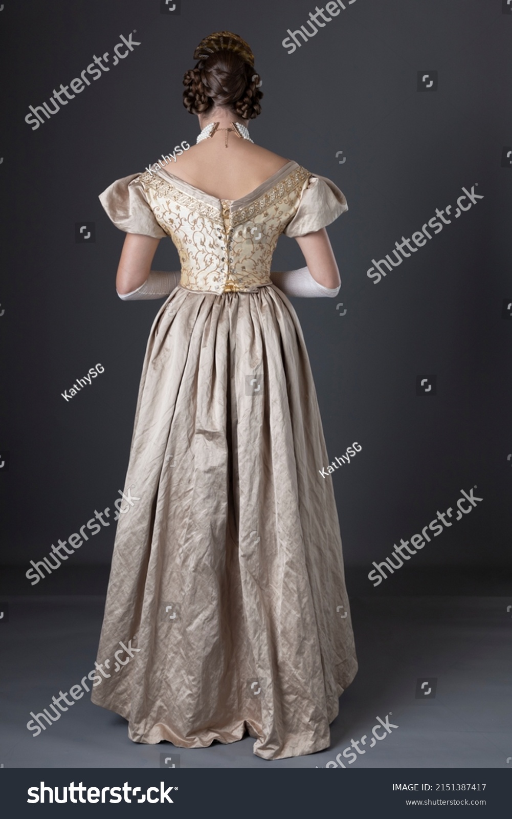 A Victorian woman wearing a gold ball gown with long, fingerless lace gloves and standing against a studio backdrop #2151387417
