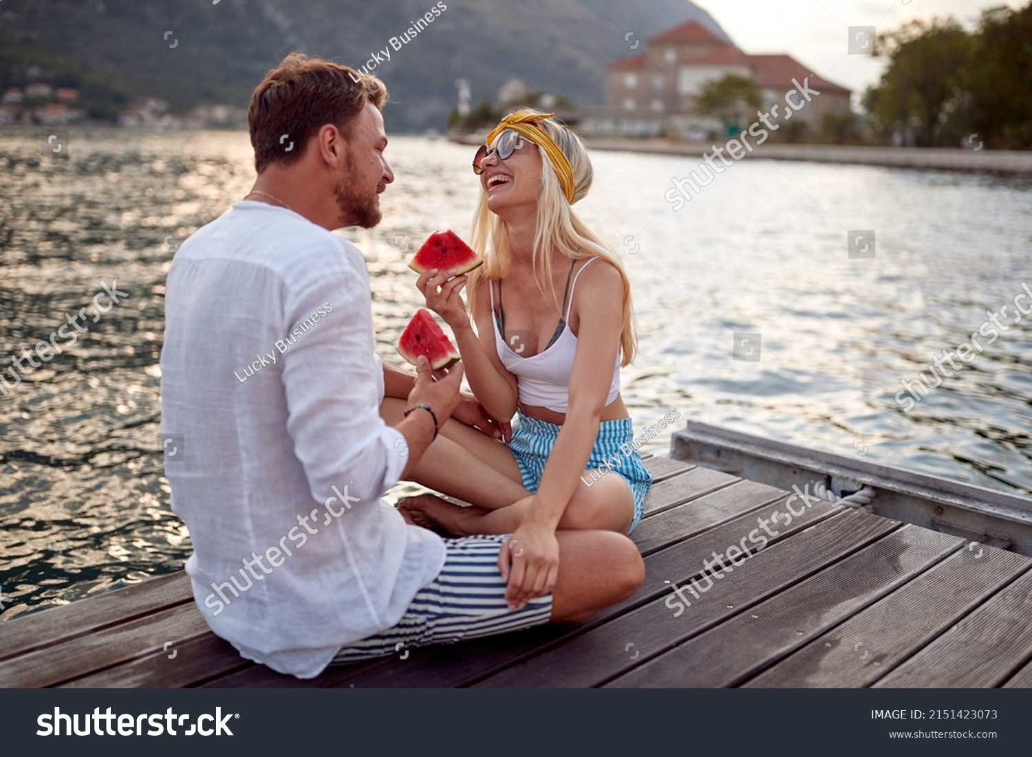 Couple having good time on date by sea siting on wooden dock, laughing and eating watermelon. Love, fun, togetherness concept. #2151423073