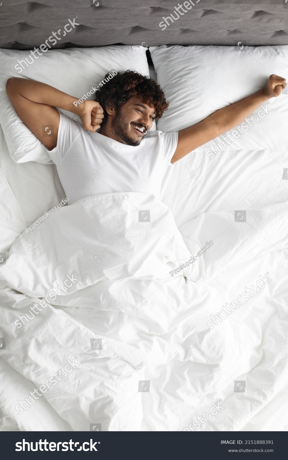 Happy curly hindu guy in white t-shirt stretching body while laying in comfortable bed, looking at copy space and smiling, enjoying new day, feeling fully rested, top view, vertical shot #2151888391