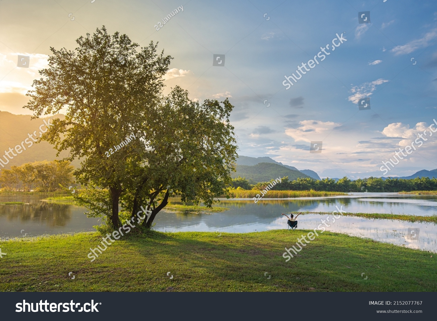 woman enjoying landscape sitting on bench at lake waiting for sunrise alone with nature and relax. Traveling and camping concept. Camping tent in forest near lake with grassland and blue sky. #2152077767