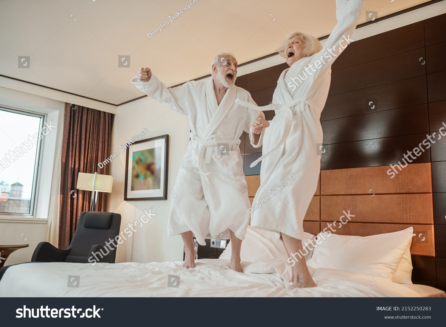 Senior couple in white robes jumping on bed and looking happy #2152250283
