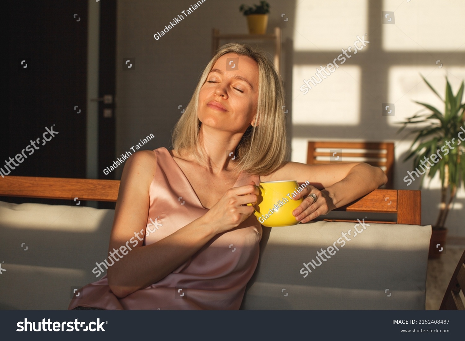 Satisfied middle aged woman is sitting on couch at home with cup of coffee. Smiling adult female of 40 years old with close eyes is sitting on sofa in living room #2152408487