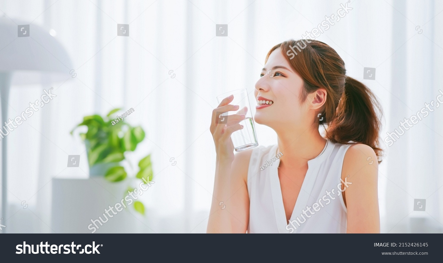 side view of asian woman with brunette ponytail smile drinks a glass water happily in a white room #2152426145