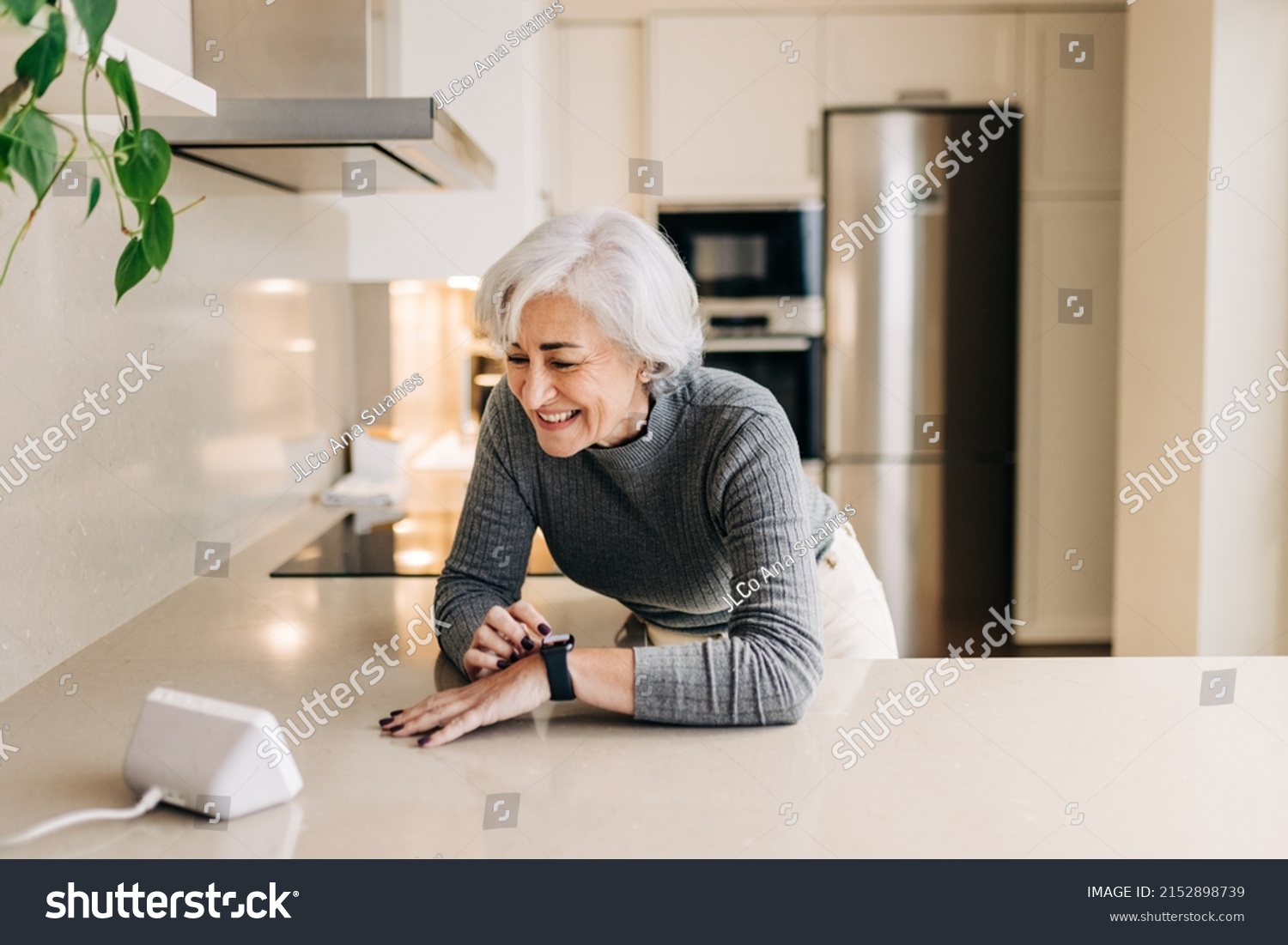 Senior woman smiling happily while using smart devices in her kitchen. Cheerful elderly woman using a home assistant to perform tasks at home. #2152898739