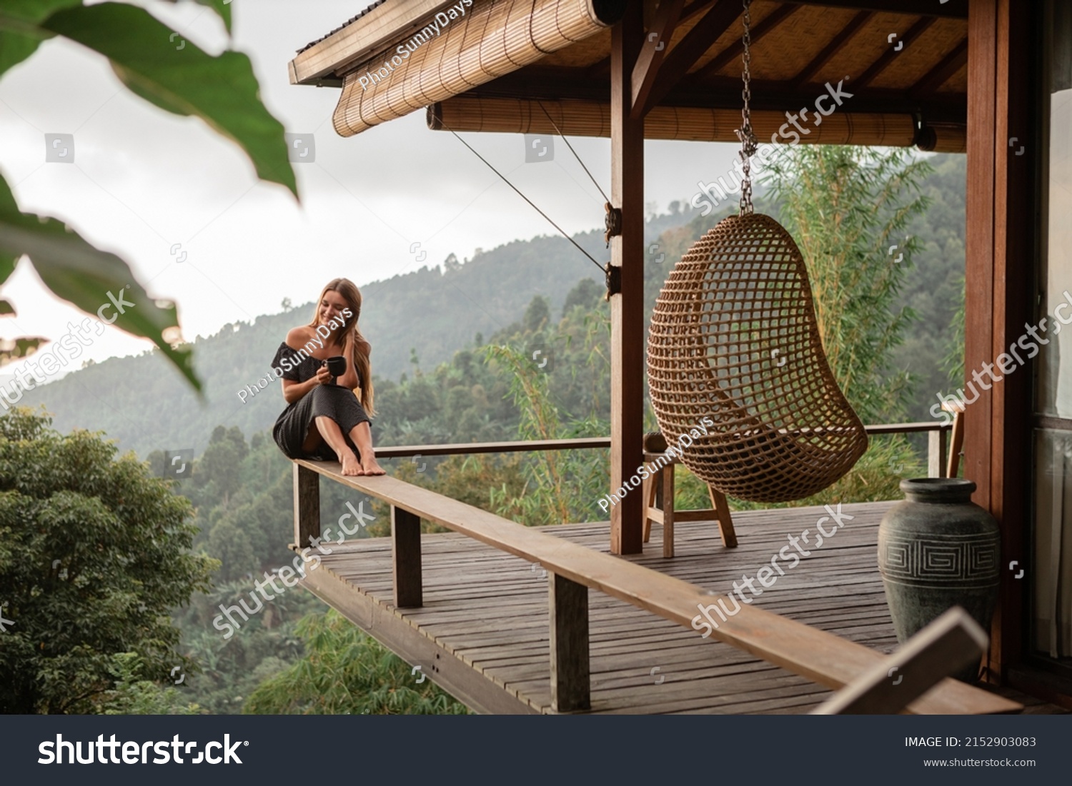 Young beautiful woman drinking coffee while sitting on a balcony in a wooden house overlooking the mountains #2152903083