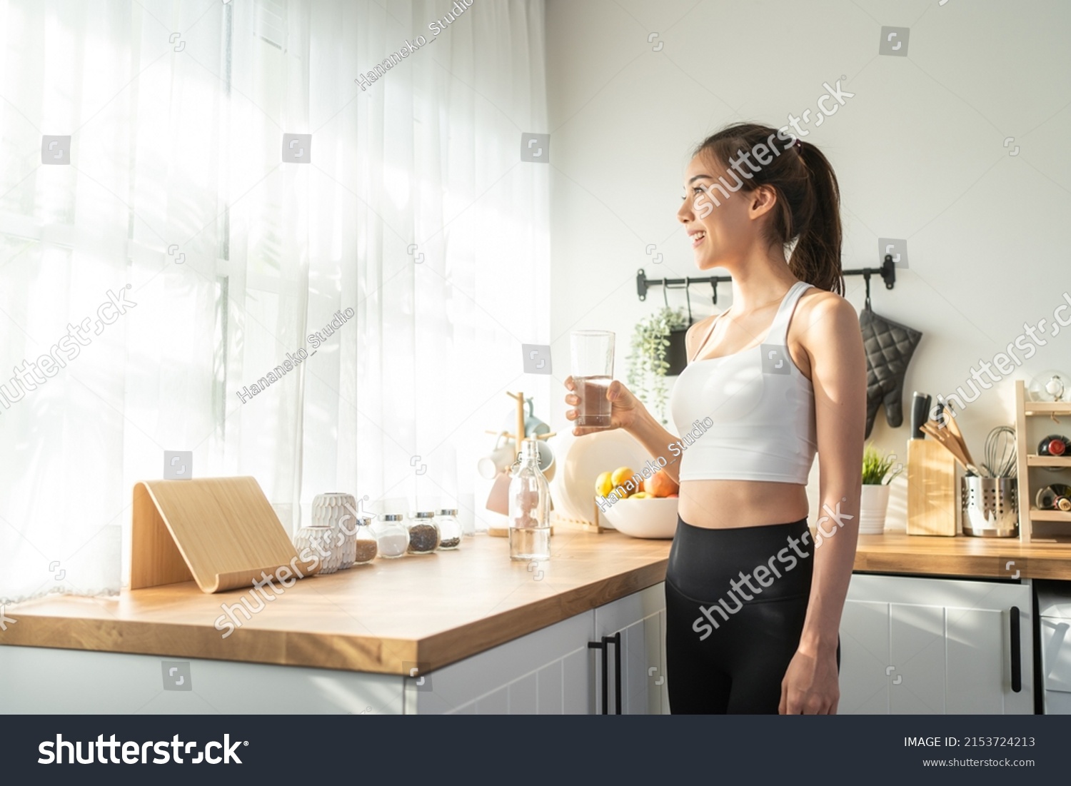Asian beautiful woman in sportswear drink water after exercise at home. Young thirsty active sport girl takes a sips of clean mineral natural in cup after workout for health care in kitchen in house. #2153724213