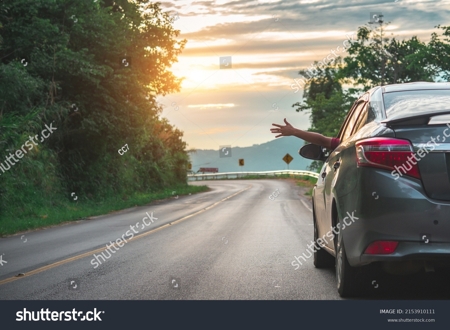 Happy woman waving hand outside open window car with meadow and mountain background. Female lifestyle relaxing as traveler on road trip in holiday vacation. Transportation and travel. #2153910111